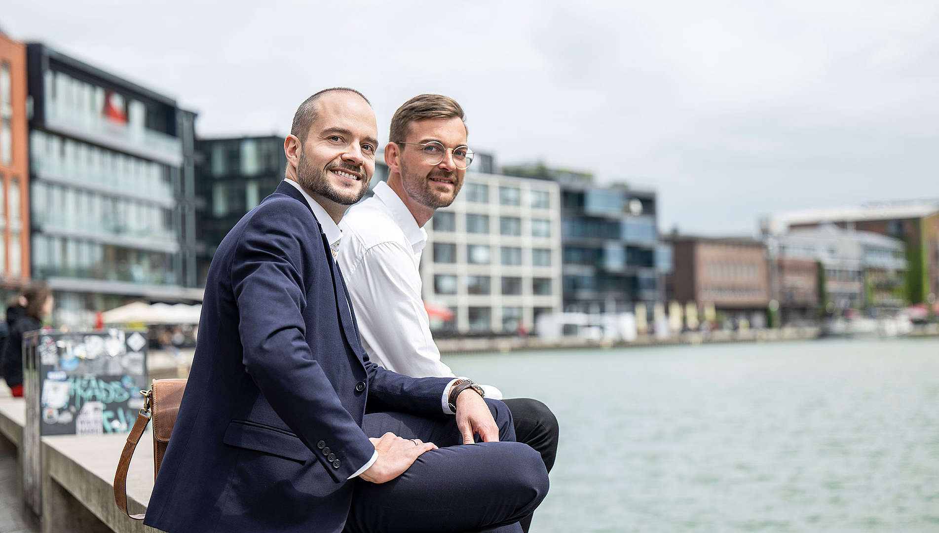 Zwei Personen sitzen am Hafen in Münster und lächeln in die Kamera.