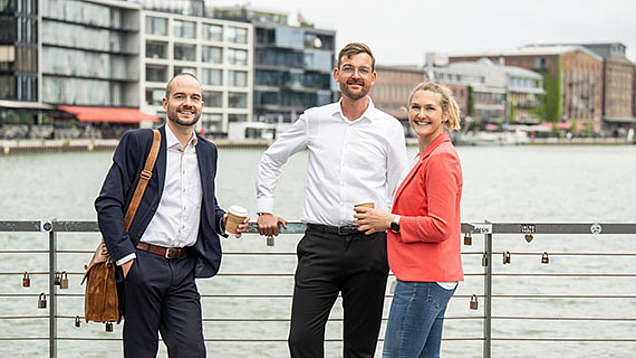 Drei Mitarbeitende, die mit einem Kaffee in der Hand am Hafen in Münster stehen und in die Kamera lächeln