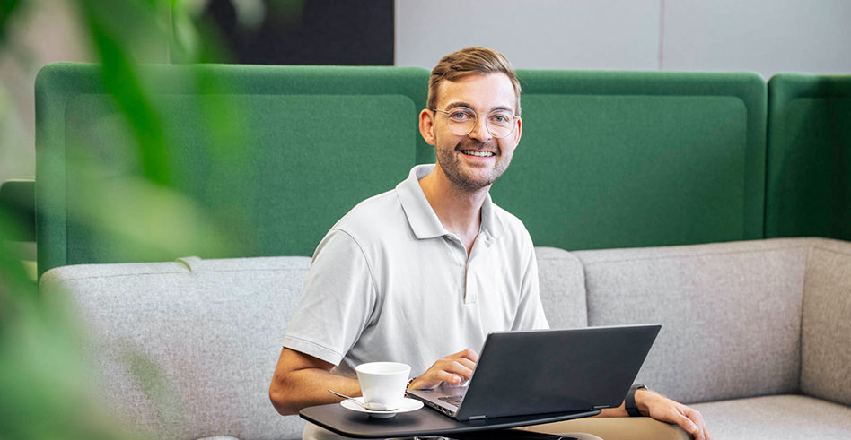 Ein Mann sitzt vor seinem Laptop und schaut fröhlich in die Kamera.